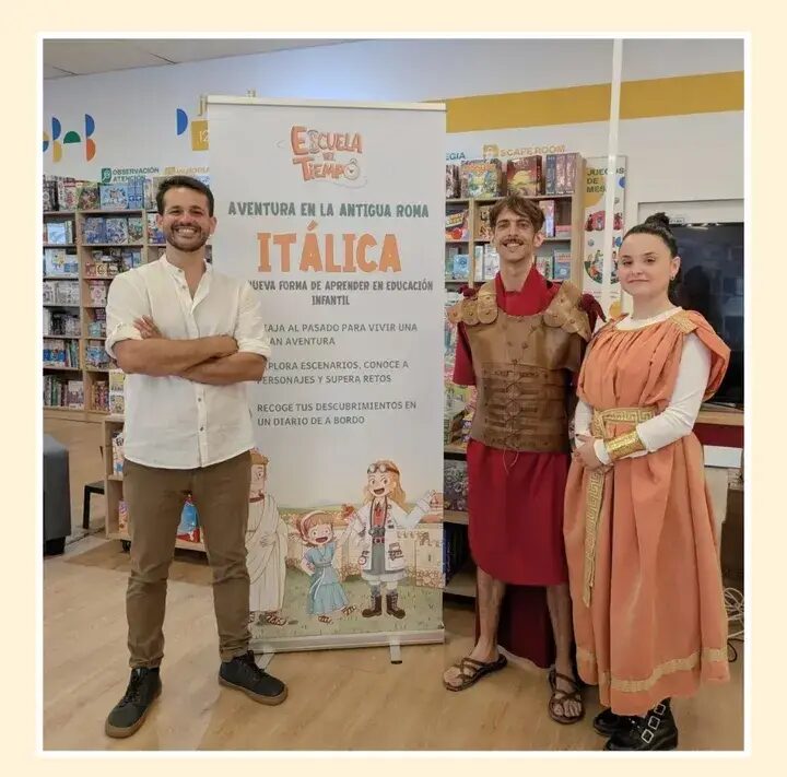 Daniel Casado y dos figurantes vestidos de romanos junto al cartel de la Aventura en la Antigua Roma: Itálica